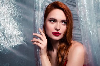 Parlak kızıl saçlı çarpıcı bir genç kadın sanatsal plastik arka planda poz veriyor, stüdyo ortamında tarzı ve zarafeti kucaklıyor. Güzellik, moda ve kadınlığın ön plana çıkarılması.