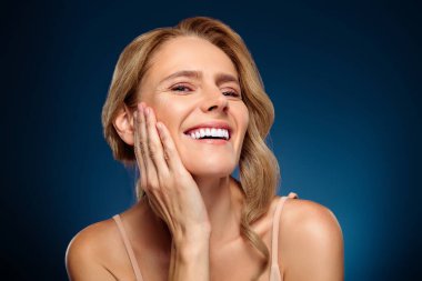 Neşeli, orta yaşlı bir kadın, pürüzsüz bir ten ve profesyonel stil ile kendine güvenen, sağlığın, güzelliğin ve kadınsı zarafetin örneklerini sunan, mavi bir stüdyo arkaplanında..