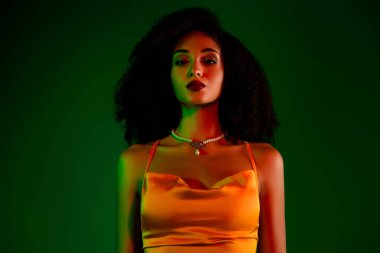 Turuncu giyinmiş genç bir kadının zarif bir görünüşü, sanatsal ışıklandırması ve görünüşünü güzelleştiren yeşil bir arkaplanı olan şık bir portresi..