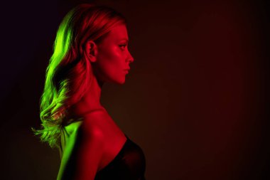 Büyüleyici genç bir kadın, neon kırmızı ve yeşil ışıklarla aydınlanmış, koyu bir arka plana karşı, güzelliği ve cazibeyi ön plana çıkaran. Zarif duruşu ve çarpıcı özellikleri şehvetli bir cazibe yayıyor..