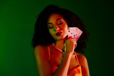 Şık bir kadın iskambil kağıtlarıyla poz veriyor zarafet ve cazibeyi somutlaştırıyor. Yeşil eğimli arka plan onun canlı, kendinden emin varlığını tamamlıyor, lüks ve sofistike bir şekilde yayılıyor..