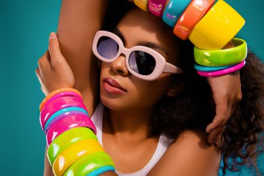 Renkli bilezikleri ve retro güneş gözlükleriyle şık bir genç kadının portresi canlı bir deniz mavisi zeminine karşı cazibe, tarz ve entelektüellik yayıyor..