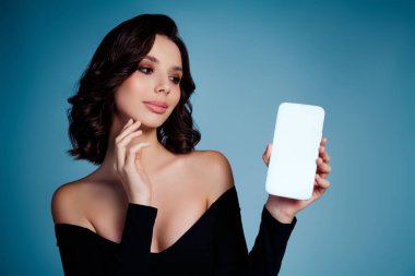 Siyah giyinmiş şık bir genç kadın, akıllı telefonuyla stüdyoda mavi bir arkaplan karşısında poz veriyor, zarif ve modaya uygun bir stil sergiliyor, büyüleyici ve modern bir çekiciliği var..