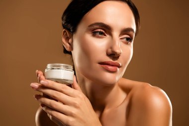 Genç bir esmer kadın bir kavanoz krema sunar, bej arkaplanda cilt bakımını ve doğal güzelliği vurgular..