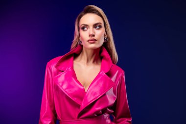Parlak pembe bir trençkot giyen göz kamaştırıcı bir kadın mor bir arka plana karşı duruyor, zarafet ve ihtişam saçıyor..