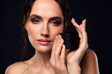 Koyu saçlı ve doğal makyajlı bir model cilt bakımı için kozmetik bir kavanoz sergiler. Zarif ve yumuşak estetik, sade bir arka plana karşı güzelliği, lüksü ve kadınsı cazibeyi vurgular..