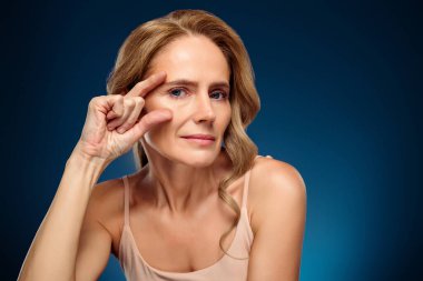 Kendine güvenen olgun bir kadın zarif bir şekilde mavi bir arka planda, güzellik ve cilt bakımını vurguluyor, dingin ifadesi ve parlak cildiyle temsil ediliyor, zarafet ve kadınsı cazibeyle..