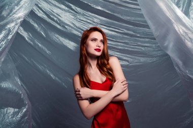 Parlak kızıl saçlı güzel bir model. Şık bir elbise içinde zarafet ve cazibe sergiliyor. Soyut bir stüdyo ortamında dokulu malzemelerle çevrili..