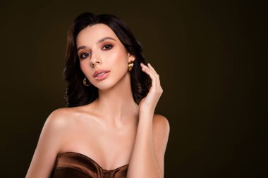 Çekici esmer kadın zarif ve şık kahverengi straplez elbisesiyle şık bir tarz sergiliyor. Onun sofistike görünüşü altın küpeler, ince makyaj ve yumuşak bukleler ile tamamlanır, koyu nötr bir zemin üzerine kurulmuş.