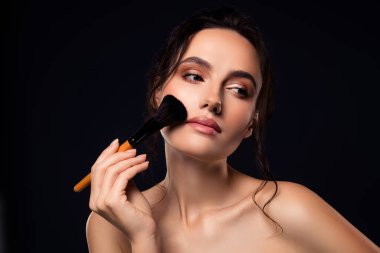 Uygulama için makyaj fırçası kullanan zarif genç bir kadın koyu bir arka planda poz veriyor, göz kamaştırıcı ve güzel.