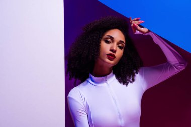 Beyaz giyimli, neon ışıklarla aydınlatılmış genç bir kadın, canlı mor ve mavi bir arka plana karşı şık bir poz veriyor..