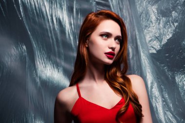 Görüntü, kırmızı saçlı ve makyajlı, kırmızı elbiseli, desenli plastik arka plan, somut stil, zarafet ve cazibeye karşı duran genç bir kadını yansıtıyor..