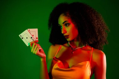 Güzel genç bir kadın, elinde canlı yeşil bir zemine karşı iki kart tutarken zarafet yayar. Kendine güveni lüks kokteyl kıyafetine uyuyor..