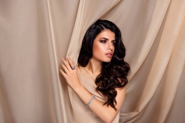 Çekici bir esmer kadın, bej kaplamalı arka planda şık moda ve lüks takılarla güzelliğini sergiliyor. Görüntü zarafeti, kadınlığı ve sofistike duyguyu yakalar..