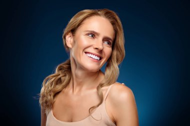 Güzel ve çekici orta yaşlı bir kadın, parlak tenli, stil sahibi sarı saçlı, ve zarif görünüşlü, canlı mavi arka planda kendine güvenen biri.