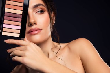 Güzel bir kadın model makyaj paleti sergilerken koyu bir arkaplanda zarif bir poz verir, kozmetik ve modern modaya vurgu yapar..