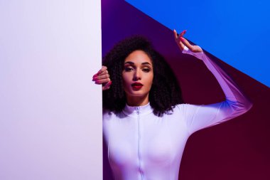 Tarz sahibi genç bir kadın dinamik bir zemin üzerinde poz veriyor, çağdaş renklerde neon ışıklarla aydınlatılıyor, modern estetik bir ortamda özgüven ve zarafet yayıyor..