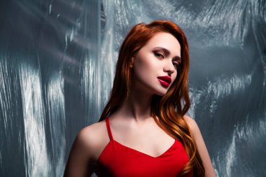 Şık kızıl saçlı, şık kırmızı elbiseli zarif bir genç bayan stüdyo ışıklarının altında sanatsal plastiğe karşı duruyor, canlı bir stil, güzellik ve çekicilik sergiliyor..