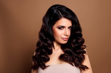 Zarif esmer kadın stil buklelerini, şık makyajını ve tarafsız bir bej stüdyo geçmişine karşı kendine güvenini gösteriyor..