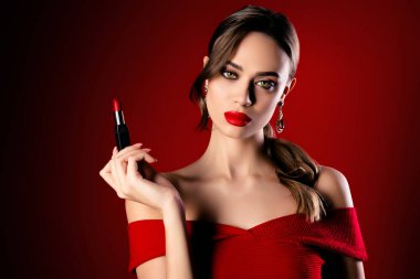 Kozmetik ürünler taşıyan, şık kırmızı elbiseli şık bir kadın, zarafeti, makyaj sanatını ve şık cazibesini vurgulayarak, şenlikli ve göz kamaştırıcı alt tonlarıyla çarpıcı bir kırmızı zemine karşı koyuyor..