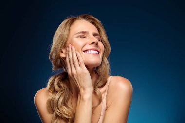 Çekici bir kadın ışıl ışıl gülüşünü ve parlak ten rengini canlı mavi bir arka plana karşı sergiliyor zarafet, moda ve profesyonel cilt bakımını sembolize ediyor..