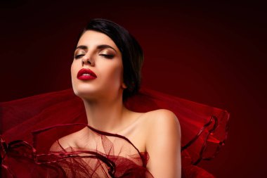 Kırmızı dudakları ve göz kamaştırıcı moda aurası olan şık bir kadının stilize edilmiş yakın çekimi, sanatsal kırmızı arka planda zarif bir kıyafet giyiyor..