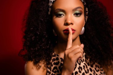 Leopar desenli bir mayo giymiş ve cesur mücevherlerle aksanlı doğal güzelliğini sergileyen ileri moda bir kadın, zengin kırmızı bir arkaplanda poz veriyor, cazibe ve retro-ilham temalar için mükemmel..