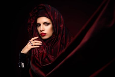 Güzel ve zarif bir kadın zarif poz verir, parlak kırmızı kumaş giyer ve koyu bir arka plan, yansıtıcı stil, sofistike ve cazibe karşısında güzel mücevherler.