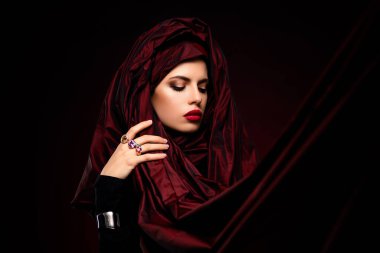 Koyu kırmızı kumaşa sarılı genç bir kadın zarif ve sofistike makyajı ve büyüleyici bakışlarıyla.