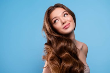 Çekici, dalgalı kahverengi saçlı, mavi bir fonda poz veren genç bir kadının yakın çekimi güzelliği ve tarzını gösteriyor.