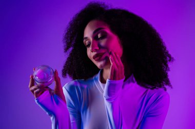 Ultraviyole neon ışıklı güzel bir kadın model kozmetik kavanozu tutuyor, güzellik, cilt bakımı ve mor ışıklı arka planda modern cazibeyi vurguluyor..