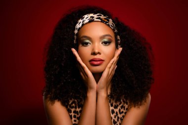 Şehvetli bukleleriyle zarif poz veren göz kamaştırıcı genç bir kadın. Leopar desenli bir elbise giymiş, şık bir saç bandı ve makyajla tamamlanmış, canlı kırmızı bir arka planı var, kendine güveni tam..