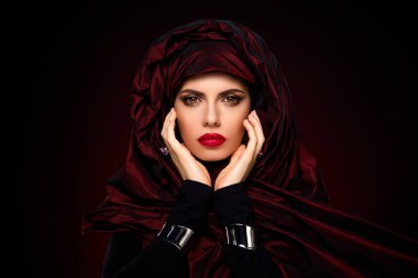 Lüks kırmızı eşarplı güzel bir genç kadının portresi, büyüleyici makyajını ve şık görünümünü vurguluyor, zengin, karanlık bir arka plana karşı şekillendirilmiş..