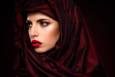 Zarif genç bir kadın zarif bir şekilde kırmızı kumaşa sarılmış poz veriyor. Kalın kırmızı dudaklar ve etkileyici gözlerle güçlendirilmiş klasik güzelliği sergiliyor. Artistik koyu renk bir arka planla çevrili..