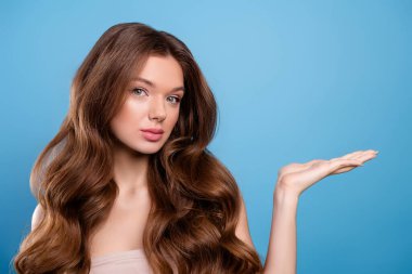 Dalgalı kahverengi saçlı ve mükemmel makyajlı genç bir kadın. Parlak mavi bir arkaplanda poz veriyor. Zarafet ve güzellik örneği..