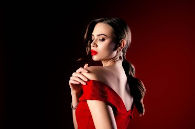 Güzel, kendine güvenen bir kadın çarpıcı kırmızı bir elbise içinde çarpıcı bir karanlık arka plana karşı zarafetle poz verir, zarafet ve entelektüellik yayar. Modaya uygun makyaj ve saç stili cazibesine vurgu yapıyor..