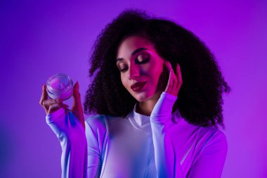 Siyah bukleli şık bir kadın koyu morötesi ışık ayarları altında bir kavanoz krem tutarak cazibe ve özgüvene vurgu yapıyor.