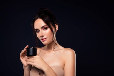Elinde kozmetik ürünler tutan genç ve güzel bir kadın. Zarafet, cilt bakımı ve moda örneği. Esmer model, lüks ve kendine güveni kucaklayan karanlık bir zemin üzerinde duruyor..