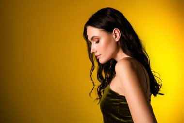 Şık, siyah saçlı, kadife yeşili elbisesi parlak sarı stüdyo arka planında poz veren şık bir genç kadın..