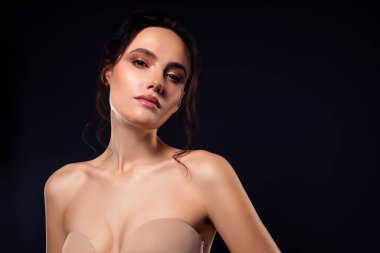 Askısız bir elbise içinde şık makyajlı esmer bir kadın karanlık bir stüdyo arkaplanına karşı poz veriyor. Güzelliğini ön plana çıkaran zarafet ve cazibe saçıyor..