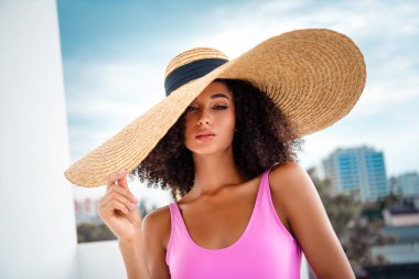 Geniş kenarlı bir şapka ve parlak pembe bir mayo giyen genç bir manken, dışarıda arka planda güneş ışığı altında zarafet ve tarz yayıyor..