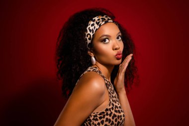 Kırmızı bir stüdyonun arka planında duran zarif genç bir kadın, şık bir leopar desenli kıyafet ve uyumlu bir saç bandı giyiyor, büyüleyici tarzını, etkileyici makyajını ve büyüleyici cazibesini vurguluyor..