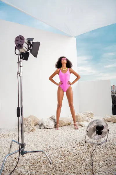 Pembe mayo giyen kendine güvenen bir kadın manken, moda çekimi için dışarıda duruyor, profesyonel ışıklarla çevrili, arka planda güzel bir yaz gökyüzü var..