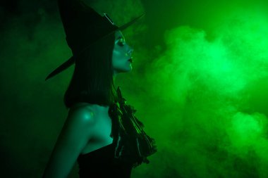 Cadı gibi giyinmiş şık bir kadın, yeşil ışıkta yıkanmış ve etrafı sis, modern cadılar bayramı modasıyla çevrili.