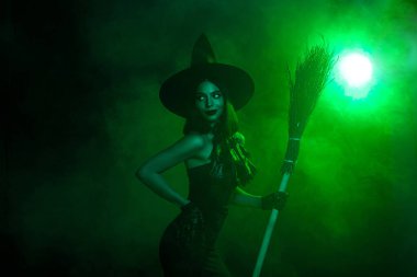 Cadı kostümü giymiş güzel bir kadın elinde yeşil sisli bir atmosferde süpürge tutuyor. Cadılar Bayramı ve fantezi temalarına uygun..