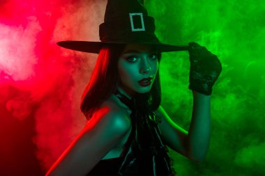 Büyüleyici bir kadın şık bir cadı şapkası takıyor dumanlı kırmızı ve yeşil ışıkta büyüleyici bir şekilde poz veriyor, mükemmel bir şekilde canlandıran fantezi ve Cadılar Bayramı ruhu..