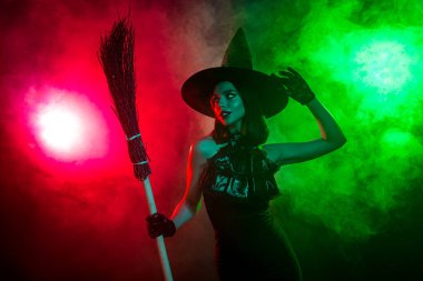 Siyah elbiseli ve şapkalı şık bir cadı elinde süpürge, dramatik ışıklandırma ve atmosferik sis ile çevrili..