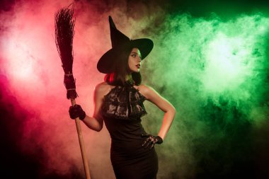 Elinde süpürge sopasıyla cadı gibi giyinmiş büyüleyici bir kadın koyu pembe ve yeşil bir sis içinde poz veriyor ve büyülü Cadılar Bayramı titreşimleri yayıyor..