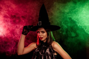 Cadı gibi giyinmiş şık bir kadın zarif bir şekilde duruyor. Etrafı yeşil ve kırmızı tonlarda gizemli dumanlı ışıklarla çevrili. Sonbaharda Cadılar Bayramı cazibesi ve büyülü bir gizem yayıyor..