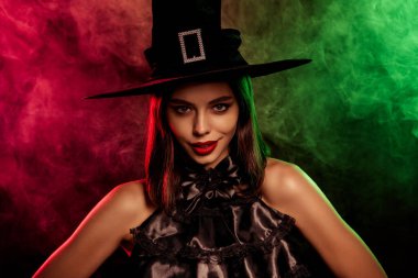 Siyah bir cadı kostümü ve şapka giyen çekici bir kadın renkli bir sisin önünde poz veriyor ve Cadılar Bayramı temalarını çağrıştırıyor..